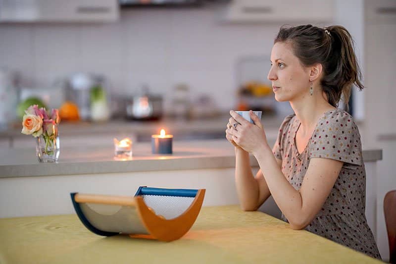 Eine junge Frau sitzt an einem Küchentisch und hält mit beiden Händen eine Teetasse fest.