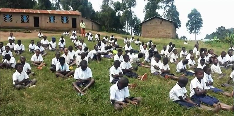 Mehrere afrikanische Schulkinder praktizieren die Transzendentale Meditation im freien auf einer grüner Wiese.