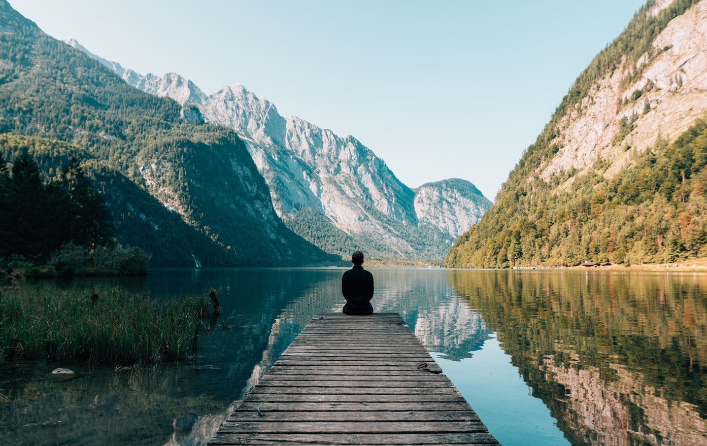 Eine Person sitzt auf einem Steg. Vor ihr ein See und Berge.