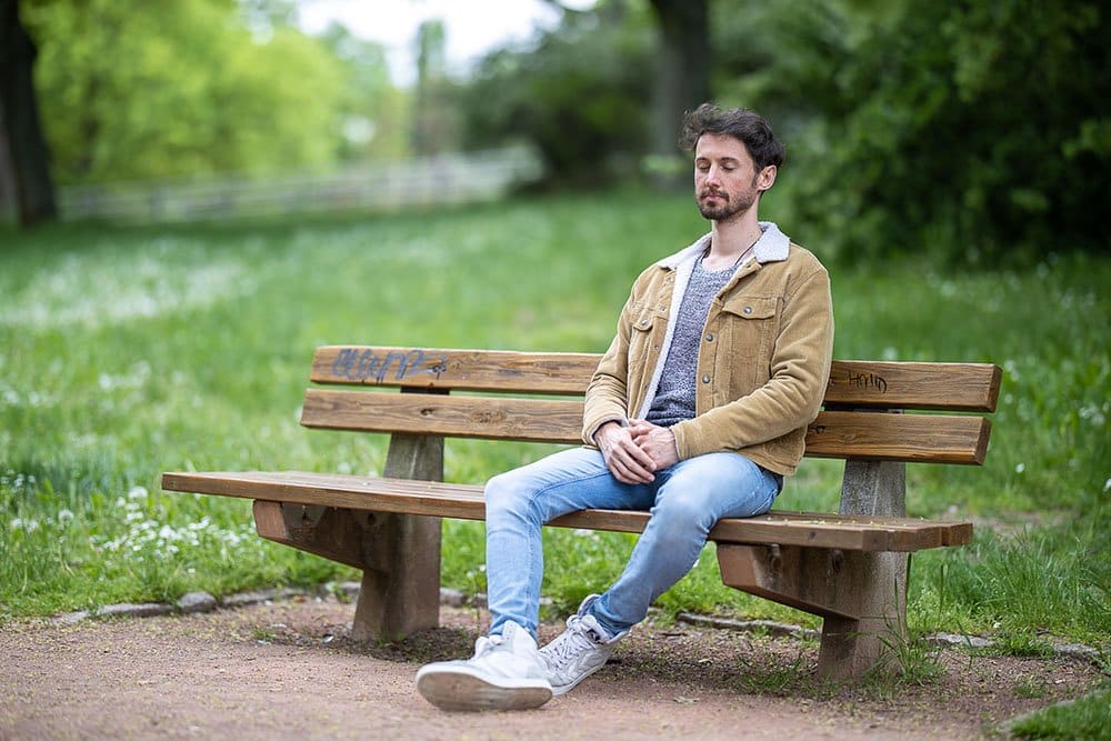 Ein junger Mann sitzt im freien auf einer Holzbank und meditiert.