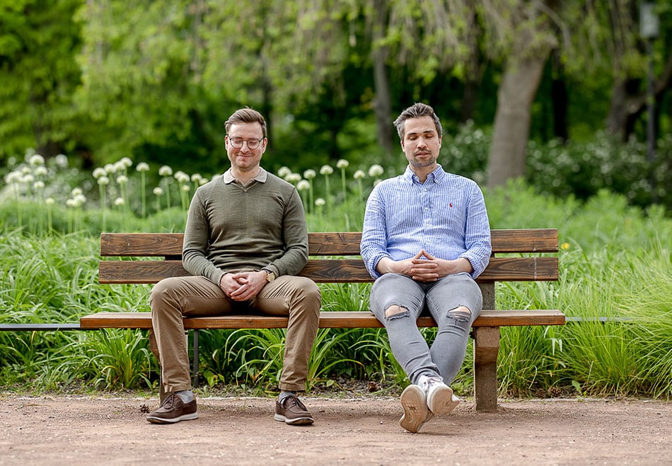 Zwei junge Männer sitzen mit geschlossenen Augen auf einer Parkbank und meditieren.