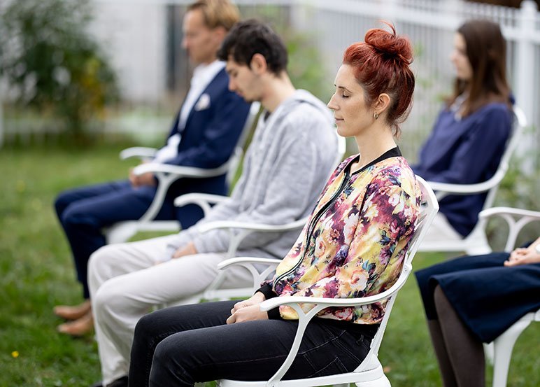 Eine Gruppe Meditierender sitzt und meditiert draußen auf Gartenstühlen.