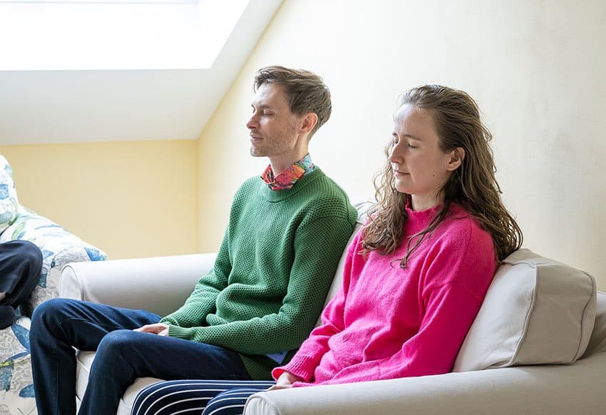 Zwei junge Personen sitzen entspannt auf einem Sofa und meditieren.