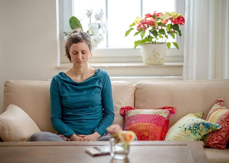 Eine junge Frau sitzt auf einem Sofa und meditiert.