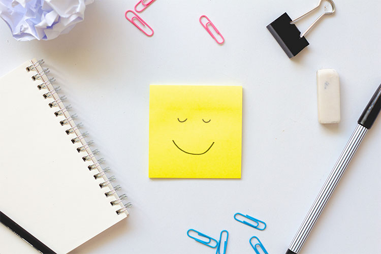 Verschiedene Schreibtischartikel. Im Vordergrund ein gelber Klebezettel mit meditierendem Smiley
